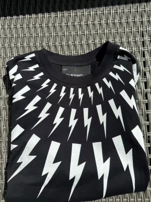 Neil Barrett svart t-shirt - Svart t-shirt från Neil Barrett med slim fit och ikoniskt blixtmönster i vitt över hela framsidan. Rund halsringning och mjukt material som känns skönt mot huden. Perfekt för dig som vill sticka ut med en grafisk och modern look. Nypris ligger på runt 4000kr.