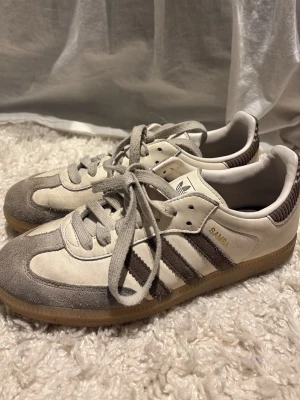 Adidas Samba vita och grå sneakers - Säljer ett par klassiska Adidas Samba sneakers i vitt och grått med bruna ränder. Skorna har ovandel i mocka och läder, gummisula och snörning. Perfekta för dig som gillar retrostil och tidlös design.