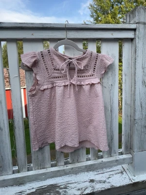 Ljusrosa blus med spetsdetaljer - Söt ljusrosa blus med korta ärmar och dekorativa spetsdetaljer upptill. Blusen har en knytning vid halsen och volangkant längs bröstet. Tillverkad i ett mjukt, strukturerat tyg som ger en romantisk känsla.🩷