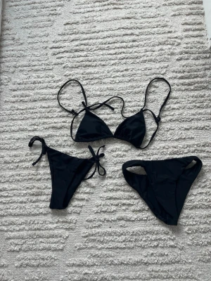 Svart bikini med trekantsöverdel - Snygg svart bikini med klassisk trekantsöverdel och två olika underdelar – en med knyt i sidan och en med bredare sidor. Perfekt för stranden eller poolen, enkel att matcha med andra plagg. Materialet är stretchigt och bekvämt.