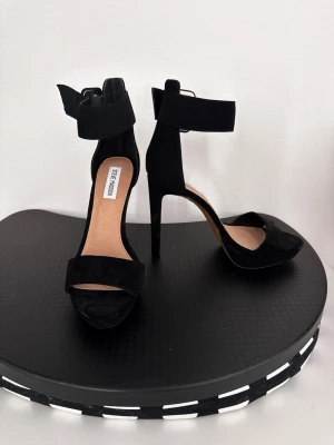 Svarta klackpumps från Steve Madden - Snygga svarta pumps med hög klack från Steve Madden. Skorna har öppen tå, bred rem över tårna och en bred ankelrem med spänne. Tillverkade i mockaliknande material och har en elegant siluett som ger en trendig look.