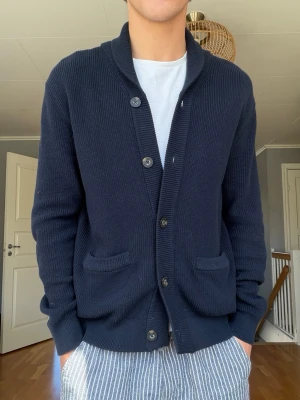Bondelid mörkblå cardigan – Storlek S - Stilren mörkblå stickad cardigan från Bondelid i storlek S. Klassisk modell med sjalkrage, knäppning framtill och två fickor. Sitter snyggt och fungerar perfekt över t-shirt eller skjorta.  Fint bruksskick med normalt mindre slitage. Passformen syns på bilderna.  