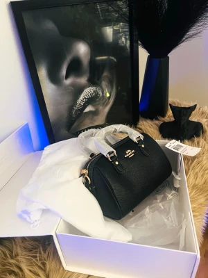 Coach-väska (Modell: Mini Rowan Satchel) - Helt ny och oanvänd Coach-väska - Väldigt elegant! Letar du efter den perfekta väskan som får dig att glänsa på festen? Då har du gjort ett utmärkt val! Säljer en helt ny, oöppnad och äkta Coach-väska (Modell: Mini Rowan Satchel) i klassisk svart färg med guldfärgade detaljer. Den ar absolut sprillans ny - skyddsplasten sitter fortfarande kvar på handtagen och prislappen hänger kvar Perfekt som en exklusiv present till dig själv eller någon du tycker om. Hör gärna av dig om du har några frågor.
