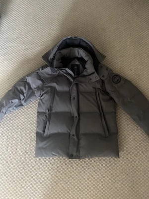 Grå pufferjacka med huva - Snygg grå pufferjacka med stor huva och quiltad design. Jackan har två sidofickor, knapp- och dragkedjestängning samt en patch på ärmen. Perfekt för kalla vinterdagar och riktigt varm tack vare det tjocka materialet. Päls kommer med