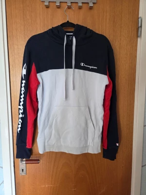 Champion hoodie i navy, grå och röd - Snygg hoodie från Champion i färgblock med navyblå upptill och på ärmar, vit framtill och röd detalj längs ärmarna. Stor Champion-logga på ena ärmen och mindre logga på andra armen. Klassisk huva med dragsko och stor magficka. Perfekt för en chill och sportig look. Har lite missfärgning (se bild 6) 