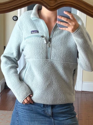 Ljusblå fleece från Patagonia - Mysig ljusblå fleece från Patagonia med halv dragkedja framtill och hög krage. Tröjan har en bröstficka med dragkedja och två sidofickor med dragkedja. Perfekt för lager-på-lager och chill dagar. I gott skick - nypris 1500kr