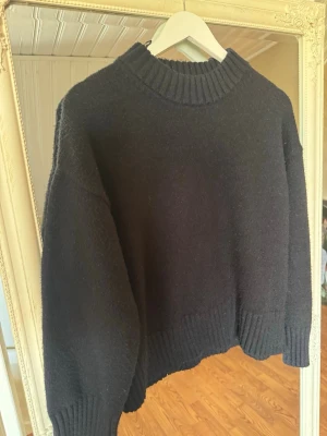 Svart stickad tröja från Gina Tricot - Mysig svart stickad tröja från Gina Tricot med hög, ribbad polokrage och breda ribbade muddar vid ärmslut och nederkant. Tröjan har en avslappnad passform och är perfekt för kyliga dagar. Materialet känns mjukt och värmande.