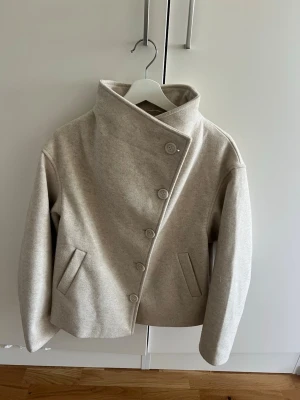 Beige kappa med knäppning - Stilren beige kappa från 157 med hög krage och snygg knäppning framtill. Kappan har två fickor och en rak passform. Perfekt för dig som gillar minimalistisk och modern stil.