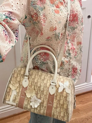Dior handväska med blommor - Säljer en handväska från Dior den heter Mini Boston Trotter med det klassiska Dior monogrammönstert och ett axelband, som jag tillverkat själv. Väskan har vita handtag, detaljer i vitt skinn, så  som dekorativa blommor och rosett och en etta i vitt skinn och med strass och namnet Dior framtill. Dior hänglås sitter på också men det kan jag skicka separata bilder på om du vill? Till dig som vill ha en unik och trendig accessoar.