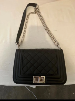 Svart quiltad axelväska med kedja - Snygg svart axelväska med quiltat mönster och silverfärgad kedjerem. Väskan har en rektangulär form och ett elegant spänne framtill i metall. Perfekt för dig som gillar stilrena accessoarer med en modern touch. Materialet ser ut att vara konstläder.