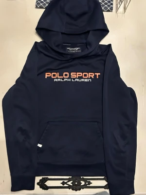 Mörkblå hoodie Polo Sport - Snygg mörkblå hoodie från Polo Sport Ralph Lauren med stor logga på bröstet och P67-tryck på ryggen. Hoodien har en klassisk huva och en stor magficka. Materialet är mjukt och bekvämt, perfekt för en avslappnad look. Perfekt för att träna med, den har en liten skavank som man ser på sista bilden. Men inget man ser vid användning! Det är storlek L i juniorer som är storlek S eller XS
