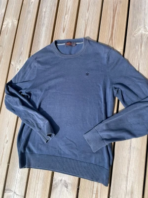Mörkblå stickad tröja från Morris - Snygg mörkblå stickad tröja från Morris med rund halsringning och diskret broderad logga på bröstet. Tröjan har ribbade muddar vid ärmslut och nederkant. Perfekt till jeans eller chinos för en clean look. 
