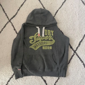 Superdry Hoodie - Säljer en grå hoodie från Superdry med tryck i gult på framsidan. Tröjan har en justerbar huva med snören och en stor ficka framtill. Perfekt för en avslappnad stil. Storleken är S men passar även XS.