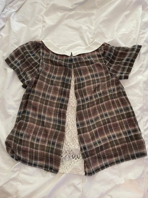 Rutig blus med spets och krage - Söt rutig blus i bruna och grå toner med vit krage och spetsdetalj nedtill. Blusen har korta ärmar och en öppen rygg med spetsinfällning som ger en unik look. Köpt på secondhand i Japan 