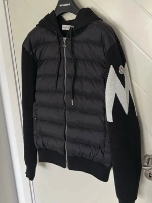 Moncler cardigan - Exklusiv Moncler cardigan med huva och dragkedja framtill. Jackan har stickade ärmar och en stor vit Moncler-logga på ena ärmen. Jackan är quiltad och vadderad för extra värme. Perfekt för kalla vinterdagar. Cardiganen är i storlek L men passar även M. Vid intresse är det bara att höra av sig. Fraktar inom 24h efter köp 📦