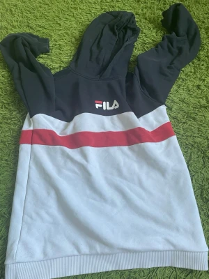 Svart och vit hoodie från FILA - Snygg hoodie från FILA i svart och vitt med röda detaljer. Stor logga på ryggen och mindre logga framtill. Hoodien har huva och långa ärmar, samt en bred röd rand över bröstet och ryggen. Perfekt för en sportig och avslappnad stil.