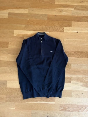 Mörkblå halvzip tröja från gant - Stilren mörkblå tröja med halv dragkedja och liten broderad logga på bröstet. Tröjan har lång ärm, ribbade muddar och en klassisk passform. Perfekt för en avslappnad men ändå snygg look.