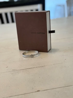 Silver armband från Louis Vuitton - Snyggt silverfärgat armband från Louis Vuitton med LV-logga framtill. Armbandet har gnistrande detaljer längs hela bandet som ger en exklusiv känsla. Perfekt accessoar för dig som gillar lyxiga smycken och vill sticka ut.