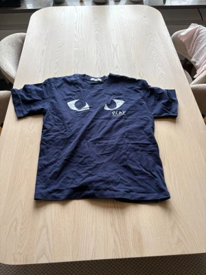 Mörkblå Comme des Garçons PLAY t-shirt - Snygg mörkblå t-shirt från Comme des Garçons PLAY med två stora ögon tryckta på bröstet och logga under. Klassisk rund hals och korta ärmar. Perfekt för dig som gillar streetwear och vill sticka ut lite extra.