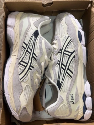 Vita och grå Asics sneakers - Snygga sneakers från Asics i vitt och ljusgrått med klassiska svarta detaljer på sidorna. Ovandelen är i mesh och mocka för en sportig look och bra andningsförmåga. Perfekta för dig som gillar stilrena och bekväma skor.