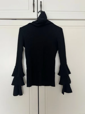 Svart stickad tröja med volangärmar - Trendig svart stickad tröja med polokrage och långa ärmar med dubbla volanger. Snygg och figurnära modell som passar perfekt till jeans eller kjol. Materialet är mjukt och stretchigt för en bekväm känsla.