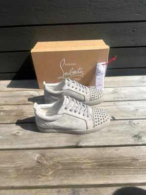 Christian Louboutin sneakers - Snygga gråa sneakers från Christian Louboutin i mocka med coola silverfärgade nitar på tån. Klassisk låg modell med matchande grå skosnören och diskret logga på hälen. Perfekta för dig som vill sticka ut med exklusiv streetstyle. Size 43, box o extra snören ingår