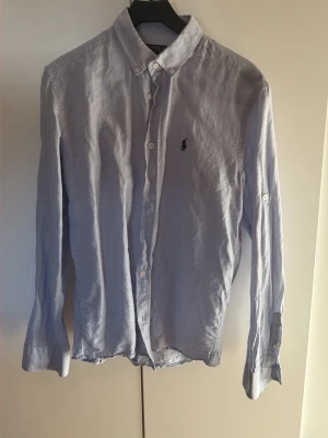 Ljusblå linneskjorta från Polo Ralph Lauren - Klassisk ljusblå skjorta från Polo Ralph Lauren i linne med button-down krage och broderad logga på bröstet. Skjortan har långa ärmar och vita knappar framtill. Perfekt för en avslappnad men stilren look. Köptes i Kroatien vet ej om äktheten.