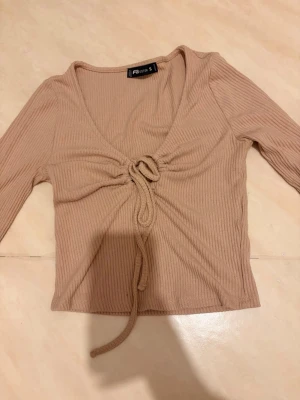 Beige ribbad topp från New Yorker - Trendig beige ribbad topp från New Yorker med lång ärm och snygg knytning framtill. Bara testad men ej använt