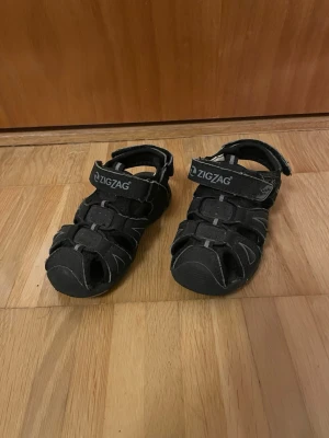 Svarta sandaler från ZigZag - Säljer ett par svarta sandaler från ZigZag med justerbara kardborreband och stängd tå. Sandalerna har vita kontrastsömmar och är tillverkade i ett slitstarkt syntetmaterial. Perfekta för sommarens äventyr eller promenader.