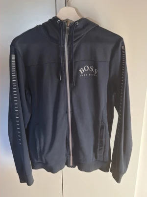 Marinblå hoodie från Hugo Boss - Snygg marinblå hoodie från Hugo Boss med dragkedja framtill och vita detaljer på ärmarna. Stor logga på bröstet och två fickor. Tillverkad i mjukt material och har huva med dragsko. Perfekt för en avslappnad och stilren look.