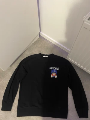 Svart sweatshirt från Moschino - Cool svart sweatshirt från Moschino med tryck av en nallebjörn och logga på bröstet. Tröjan har rund halsringning och långa ärmar. Perfekt för dig som gillar streetwear och vill ha något som sticker ut.