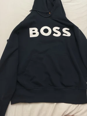 Svart hoodie med BOSS-tryck - Snygg svart hoodie från BOSS med stor vit logga på bröstet. Klassisk känguruficka framtill och huva med dragsko. Perfekt för en avslappnad och trendig look. Dessutom är den också helt ny använd 1 gång. Köpte den för 1700kr. Storlek L