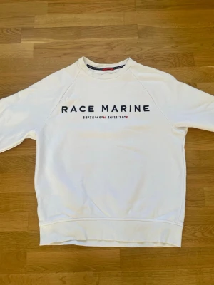 Vit sweatshirt från Race Marine - Klassisk vit sweatshirt från Race Marine med tryck i marinblått framtill och koordinater under loggan. Tröjan har rund halsringning, ribbade muddar och är tillverkad i mjukt bomullsmaterial. Perfekt för en sportig och avslappnad stil. Ej använd.