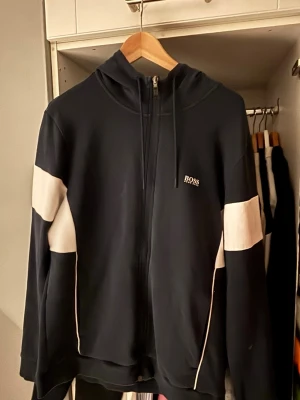 Svart hoodie med dragkedja från BOSS - Snygg mörkblå hoodie från BOSS med vit logga på bröstet och vita paneler längs ärmarna. Hoodien har dragkedja framtill, huva med dragsko och ribbade muddar. Perfekt för en avslappnad och sportig stil.