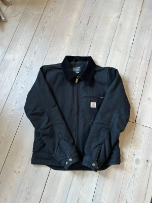Svart jacka från Carhartt - Stilren svart jacka från Carhartt med klassisk krage i manchestertyg, dragkedja framtill och bröstficka med Carhartt-logga. Tillverkad i slitstarkt material och har knappdetaljer vid ärmsluten. Funkar som både höstjackan och vinterjacka. Har använts upp till -10 grader.