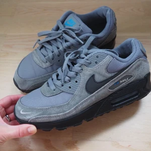 Nike Air Max 90 grå sneakers - Sonen har vuxit ur sina skor. Använda lite då och då men blev tyvärr snabbt små. Nike Air Max 90 sneakers i olika nyanser av grått med svart sula och swoosh. Ovandelen är i en mix av mocka och mesh för både stil och komfort. Klassisk Air Max-dämpning och blå logga på plösen. Perfekta för dig som gillar streetwear. Ena hälen på ena skon är lite nertrampad men inget som sonen stört sig på. Men vill att det skall stå med så att man vet! 