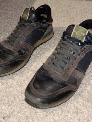 Valentino svarta och grå sneakers - Säljer ett par snygga sneakers från Valentino i svart och grå mocka och läder med mörkblå detaljer. Skorna har klassisk låg profil, snörning och en beige sula. Perfekta för dig som gillar streetwear och vill ha något unikt på fötterna.