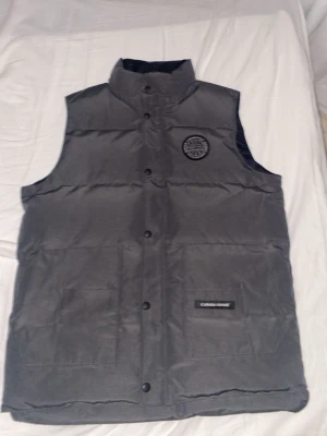 Grå dunväst från Canada Goose - Snygg grå dunväst från Canada Goose med hög krage och tryckknappar framtill. Västen har ett broderat märke på bröstet och en diskret logotyp på fickan. Perfekt för lager-på-lager och håller dig varm under kyliga dagar.