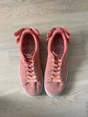 Puma sneakers med rosett - Snygga orangea/rosa Puma sneakers i ett mockaliknande material med stora satinrosetter baktill och matchande band som skosnören🩷🧡 Skorna har vit sula och klassisk Puma-logga i guld på sidan. Jättecool detalj i en outfit! Köpta på en outlet i London.