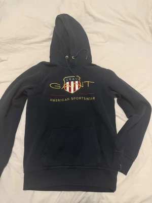 Mörkblå hoodie från GANT - Säljer en mörkblå hoodie från GANT med broderad logga och texten 'American Sportswear' på bröstet. Hoodien har huva med dragsko, känguruficka och ribbade muddar. Klassisk college-stil med snyggt broderat märke i rött, vitt och guld.