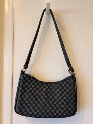 Svart axelväska med monogrammönster - Se skavankerna på remmen. Snygg svart axelväska med grått monogrammönster och justerbar axelrem. Väskan har en klassisk, rektangulär form och stängs med dragkedja. Materialet är  konstläder med detaljer i metall. Perfekt för dig som gillar stilrena accessoarer.