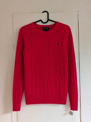 Röd kabelstickad tröja från Polo Ralph Lauren ❤️ - Klassisk cable knit-modell med svart broderad Polo-logga på bröstet. Ribbad hals, ärmslut och nederkant. En tidlös Ralph Lauren-modell som är snygg både till vardags och mer uppklätt.  Storlek L (14–16), junior.  Fint begagnat skick utan defekter!  🏷️ Polo Ralph Lauren 📏 L (14–16), junior 🎨 Röd med svart broderad logga 🧶 Kabelstickad / Cable Knit