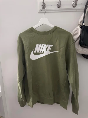 Olivgrön sweatshirt från Nike - Snygg olivgrön sweatshirt från Nike med klassisk vit logga framtill. Tröjan har rund halsringning, långa ärmar och ribbade muddar vid ärmslut och nederkant. Perfekt för en avslappnad och sportig stil. 