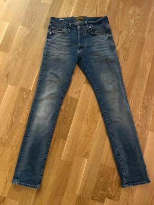 Blå slim fit jeans från Jack & Jones - Snygga blå slim fit jeans från Jack & Jones med klassisk femficksdesign och lätt slitna detaljer. Jeansen har normal midja och smala ben, perfekta till en avslappnad stil. Tillverkade i mjukt denimtyg för skön känsla. 28/32