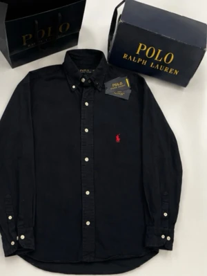 Svart skjorta från Polo Ralph Lauren - Klassisk svart skjorta från Polo Ralph Lauren med knappslå och vit knappdetalj. Skjortan har lång ärm, button-down krage och den ikoniska röda logobrodyr på bröstet. Perfekt för en stilren look.Storlek M i herr  och den är marinblå 
