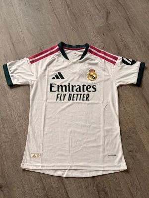 Real Madrid matchtröja Adidas vit  - ÄKTA!Snygg vit Real Madrid matchtröja från Adidas med klubbmärke på bröstet och svarta detaljer. Tröjan har rosa ränder på axlarna, mörkgröna muddar och krage samt sponsortryck på framsidan. Tillverkad i lätt och ventilerande material.
