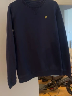 Mörkblå sweatshirt med logga - En stilren mörkblå sweatshirt med rund hals och lång ärm. Tröjan har en liten gul broderad logga på bröstet och ribbade muddar vid ärmslut och nederkant. Perfekt för en avslappnad look.
