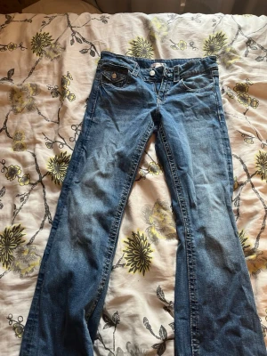Blå bootcut jeans med fickdetaljer - Snygga blå bootcut jeans med klassisk femficksdesign och dekorativa ficklock bak. Jeansen har en låg midja och är tillverkade i denim med lätt slitning framtill. Perfekta för en avslappnad och trendig look.