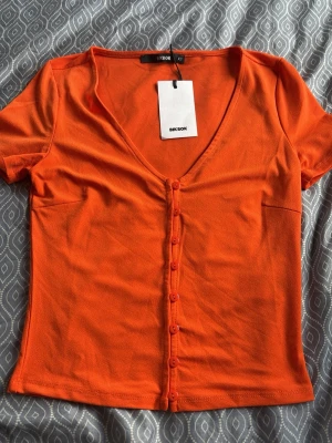 Orange kortärmad topp från Bik Bok - Jag har andra storlekar,XS,S,M,L. Snygg orange topp från Bik Bok med v-ringning och korta ärmar. Toppen har knappar framtill och är i en mjuk, stretchig bomullsblandning. Perfekt till jeans eller kjol för en färgstark look.