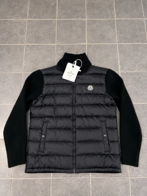 Svart pufferjacka från Moncler - Snygg svart pufferjacka från Moncler med quiltad front och stickade ärmar. Jackan har hög krage, dragkedja framtill och två fickor med knappstängning. Klassisk Moncler-logga på bröstet och stilren design som passar till allt.